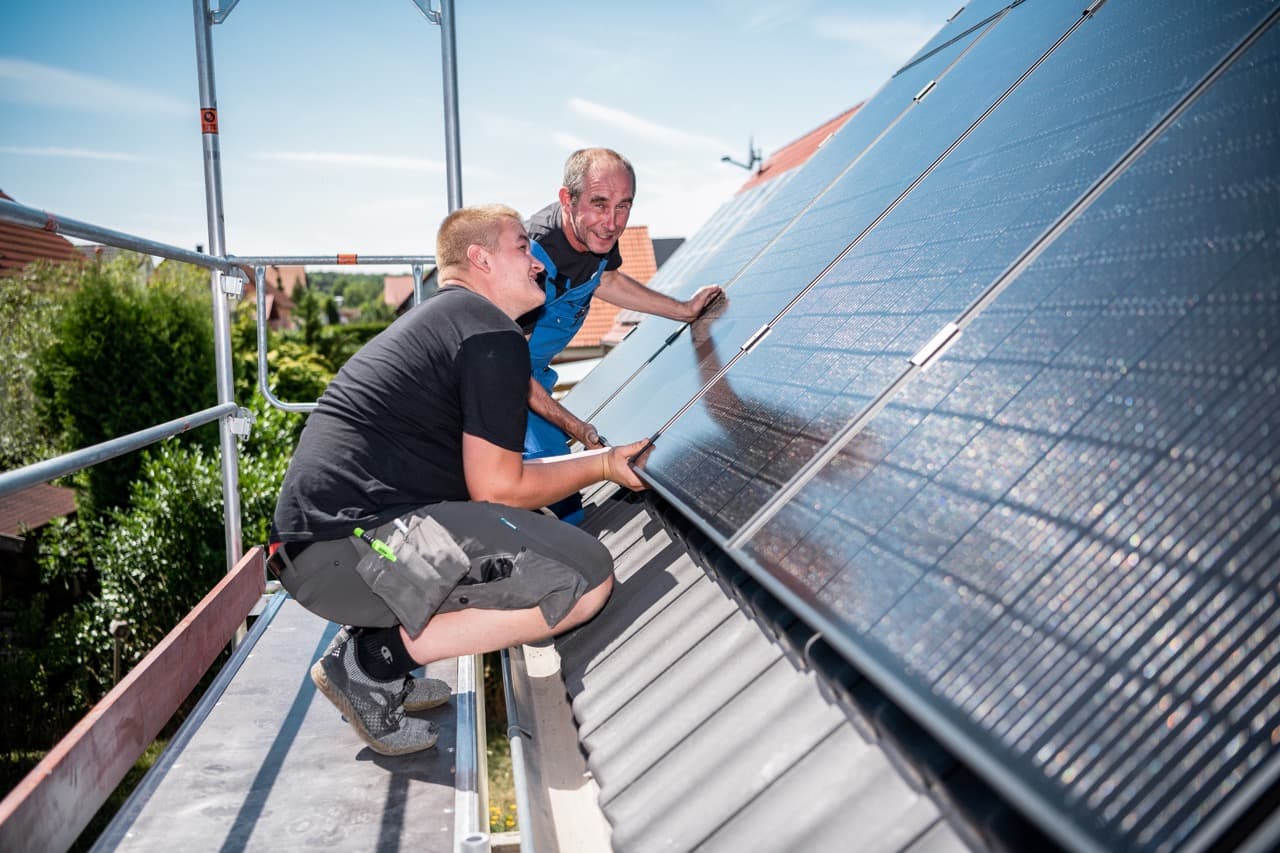 Ett tegelhus med solpaneler på taket, framför står ett stort grönt träd och i dörröppningen en liten pojke.